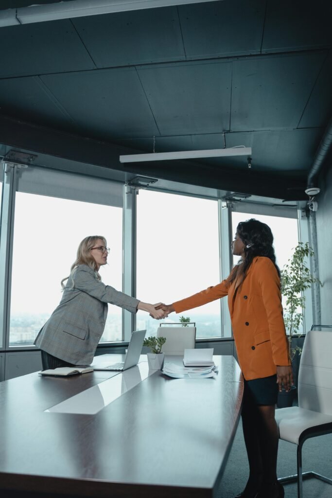Two businesswomen shake hands across a conference table in a modern office setting.